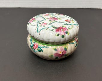 May include: A small, white ceramic trinket box with a floral design. The lid is decorated with pink and green flowers and a gold trim. The bottom of the box is decorated with pink flowers and green leaves.