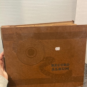 May include: A vintage brown record album with a decorative design of a vinyl record and musical notes. The words "RECORD ALBUM" are printed in bold, dark letters. The album is rectangular and appears to be made of cardboard.