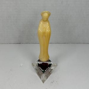 May include: A small, ivory-colored statue of the Virgin Mary with a halo, standing on a clear triangular base with a red rose.