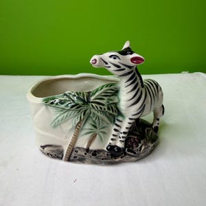 May include: A ceramic planter featuring a zebra figurine. The planter is off-white with a palm tree design. The zebra has black and white stripes, with red accents on its ears and nose. The planter is designed to hold small plants or flowers.