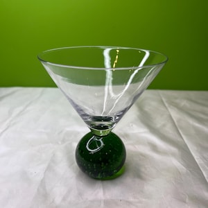 以下が含まれることがあります： 円錐形のボウルと濃い緑色の球形のベースを備えた、透明なガラスのマティーニグラス。ベースには小さな気泡があります。グラスは、緑色の背景に対して白い表面に置かれています。