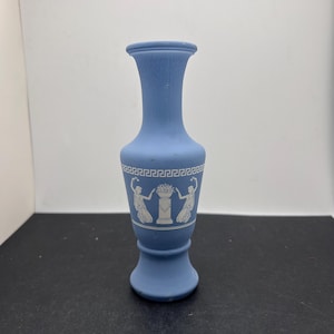 May include: A small, blue vase with white Greek-style figures and a decorative border. The vase has a flared base, a slender neck, and a rounded top. The vase is approximately 6 inches tall.