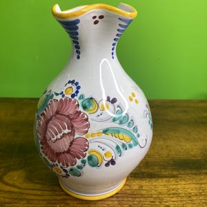 May include: A white ceramic vase with a bulbous body and a ruffled rim. The vase features hand-painted floral motifs in shades of dusty rose, teal, and yellow, with blue accents. A yellow band encircles the base and the top edge of the rim.