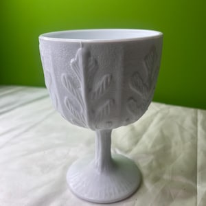 Peut inclure: Un gobelet clair avec une surface texturée et un motif de feuilles. Le gobelet a un large bol et une fine tige, reposant sur une base ronde. Le fond est vert uni.
