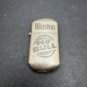 May include: A rectangular, brushed metal lighter with a hinged lid. The front surface features the engraved text Winston at the top and the words No Bull centered within a circular design. The metal has a weathered, matte finish with visible texture.