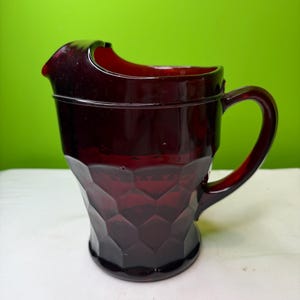 May include: A ruby red glass pitcher with a geometric pattern on the body and a curved handle. The pitcher has a spout for easy pouring. The background is a solid green color.