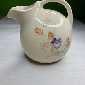 May include: A white ceramic pitcher with a floral design featuring orange, purple, and green tulips. The pitcher has a round body and a curved handle.