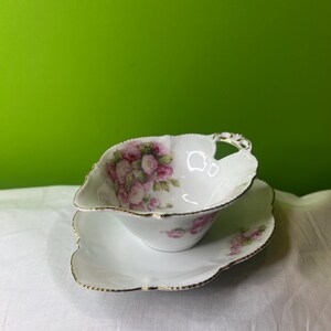 May include: A white porcelain gravy boat with a pink floral design and gold trim. The boat is on a matching saucer.