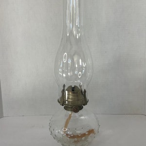 Puede incluir: Una lámpara de aceite de vidrio transparente con una base redonda texturizada y una chimenea de vidrio transparente. La lámpara tiene un quemador de latón y una mecha.