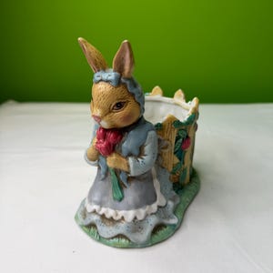 May include: A ceramic Easter bunny figurine wearing a blue dress and bonnet, holding a bouquet of red flowers. The bunny is standing in front of a small, decorative planter with a wooden fence design. The figurine has a detailed, hand-painted appearance.