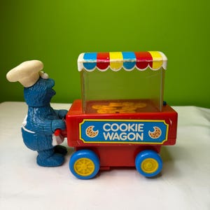 May include: A vintage Cookie Monster toy pushing a red Cookie Wagon. The wagon has blue wheels with yellow centers and a striped awning. The wagon reads "COOKIE WAGON" with cookie graphics.