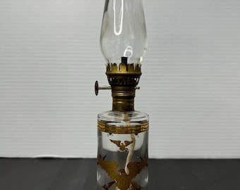 May include: A clear glass oil lamp with a gold eagle design. The lamp has a round base and a clear glass chimney.