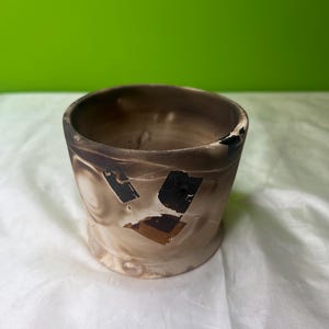 May include: A handmade ceramic cup with a unique, distressed appearance. The cup features a cylindrical shape with a slightly flared rim. The exterior showcases a blend of brown and beige tones, with dark, charred accents and geometric patterns.