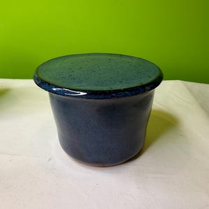Puede incluir: Una mantequera de cerámica hecha a mano con tapa. El plato es de color azul oscuro con un esmalte moteado, y la tapa es de un tono azul más oscuro. El plato tiene forma cilíndrica y está sobre una superficie blanca. El fondo es verde liso.