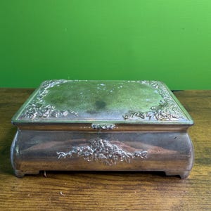 Puede incluir: Una caja rectangular de metal plateado con tapa abatible. La tapa y el panel frontal presentan motivos florales en relieve en las esquinas y en el centro. La superficie del metal muestra signos de deslustre y desgaste con un acabado liso.