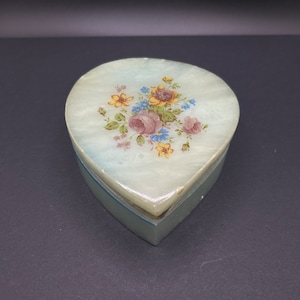 May include: A heart-shaped alabaster trinket box with a floral design painted on the lid. The flowers are yellow, pink, blue, and green.