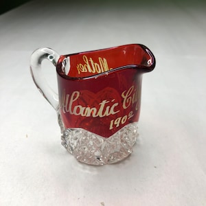 Puede incluir: Un pequeño creamer de vidrio transparente con una tapa de vidrio rojo. El creamer tiene un diseño de vidrio cortado y está decorado con letras doradas que dicen "Atlantic City 1902".