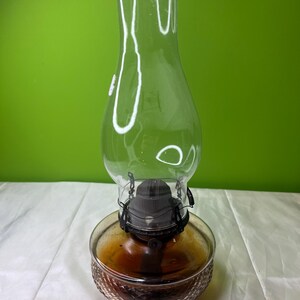 Puede incluir: Una lámpara de aceite de vidrio transparente con un depósito marrón y una chimenea de vidrio transparente. La lámpara tiene un conjunto de quemador de metal negro y una base decorativa texturizada. La lámpara está diseñada para usar con aceite.