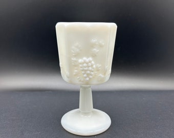 May include: A white milk glass goblet with a grape design. The goblet has a round base and a stem.