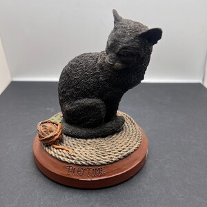 May include: A black cat figurine sitting on a round, rope-textured base with a brown rim. A small ball of twine sits next to the cat. The word "PLAYTIME" is inscribed on the base.