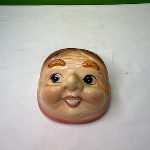 May include: A ceramic face with a smiling expression. The face has a light skin tone, with rosy cheeks and a pink outline. The eyes are blue with long eyelashes, and the hair is light brown.