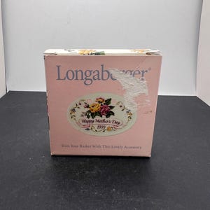 May include: A pink Longaberger box with floral accents and the text "Happy Mother's Day 1999." The box also has the text "Trim Your Basket With This Lovely Accessory."