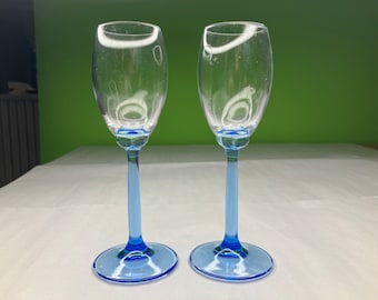 Puede incluir: Dos copas de vino de vidrio transparente con tallos y bases azules. Las copas están vacías y están sobre una superficie blanca.