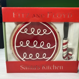 May include: A red and white ceramic Christmas ornament shaped cheese plate and spreader set by Fitz and Floyd. The set is packaged in a red box with the words "Santa's Kitchen" printed on the front.