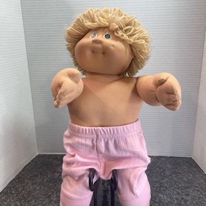 Peut inclure: Une poupée avec un visage rond en tissu beige, des yeux verts et des cheveux courts en laine blonde bouclée. La poupée a un corps souple et rembourré et porte un pantalon en tricot côtelé rose clair avec des chevilles resserrées.