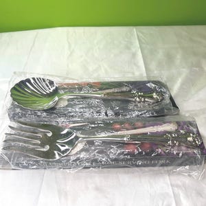 Puede incluir: Dos cubiertos, una cuchara de servir y un tenedor de servir, están empaquetados en plástico transparente. La cuchara tiene un cuenco en forma de concha. El tenedor está etiquetado como "LARGE SERVING FORK". Los mangos tienen un diseño floral decorativo.