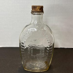 Puede incluir: Una botella de vidrio transparente con un tapón de corcho. La botella tiene un diseño texturizado y una imagen en relieve de una casa con una bandera en la parte superior.