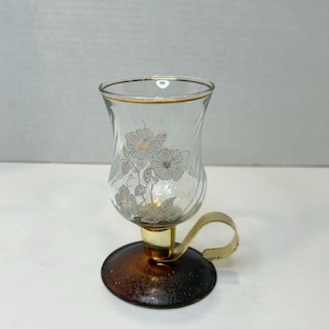 Könnte beinhalten: Ein klarer Glas-Kerzenhalter mit floralem Design und goldenem Rand. Der Halter hat eine braune Metallbasis mit goldenem Griff.