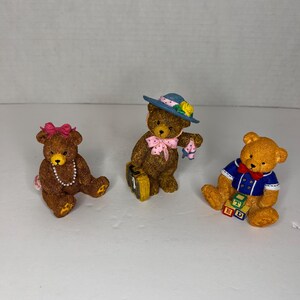 May include: Three brown teddy bear figurines. The bear on the left is wearing a pink bow and a pearl necklace. The bear in the middle is wearing a blue hat with white polka dots and a pink bow. The bear on the right is wearing a blue sailor shirt with a red bow tie and is holding colorful blocks that spell "BED".