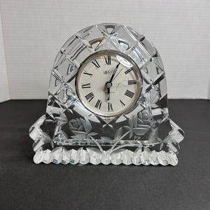 Puede incluir: Un reloj de cristal transparente con una esfera blanca y marcadores de hora romanos negros. El reloj está grabado con un diseño floral y tiene la palabra "Mikasa" en la esfera.