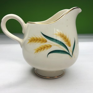 Peut inclure: Un pot à crème en céramique blanche avec un bord doré et un motif d'épis de blé jaunes et verts.