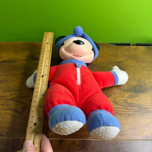 May include: A plush Mickey Mouse doll wearing a red jumpsuit with a white zipper, blue shoes, and a blue hat. The doll is approximately 12 inches tall. The doll's face is visible, with a smiling expression.