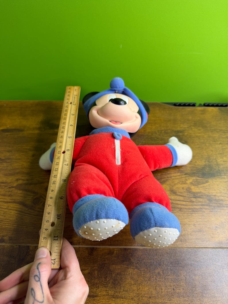 May include: A plush Mickey Mouse doll wearing a red jumpsuit with a white zipper, blue shoes, and a blue hat. The doll is approximately 12 inches tall. The doll's face is visible, with a smiling expression.