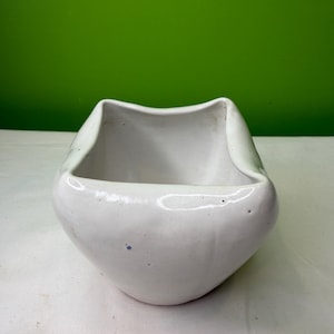 May include: A white ceramic bowl with a unique, curved design. The bowl has a square shape with rounded edges and a smooth, glossy finish. The interior is open, and the bowl is set against a green background.