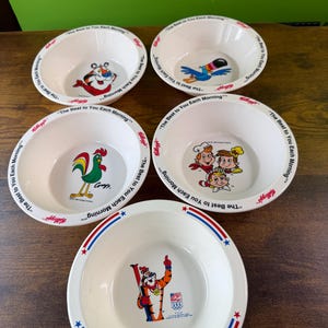 May include: A collection of five white ceramic cereal bowls. Each bowl features a different Kellogg's cartoon character in the center. The bowls have a red, white, and blue design with the text "The Best to You Each Morning".