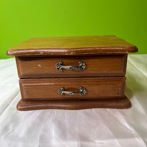 Puede incluir: Una caja de joyería de madera con dos cajones y tiradores de metal ornamentados. La caja es de color marrón claro y tiene una base ligeramente curvada.