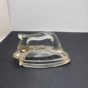 May include: A clear glass miniature iron figurine featuring a smooth, curved handle and a pointed front. The object is molded into the shape of a traditional flat iron with a solid base and a hollowed-out handle section, showcasing a transparent finish.