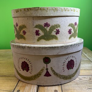 May include: Two antique, round, lidded boxes stacked on top of each other. The boxes are off-white with green and maroon floral designs. The boxes have a distressed, aged appearance, suggesting they are vintage or handmade.
