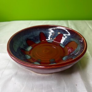 Puede incluir: Un cuenco de cerámica hecho a mano con un diseño de sol. El cuenco presenta un exterior rojo intenso, que transiciona a un interior azul y verde con detalles rojos. El centro del cuenco es de color marrón cálido.