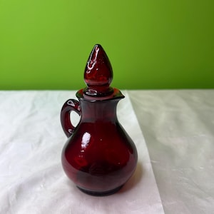 May include: A small, ruby-red glass pitcher with a handle and a pointed stopper. The pitcher has a rounded body and a small spout. The background is a solid green color.