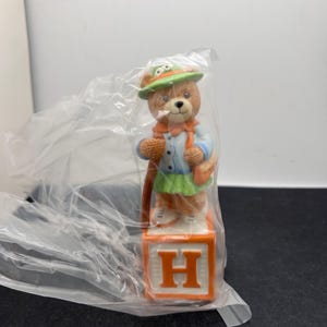 May include: A brown bear figurine wearing a green hat, orange scarf, and blue jacket, holding a satchel. The bear stands on an orange block with the letter "H" in white. The figurine is inside a clear plastic bag.