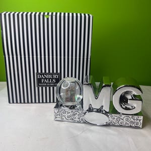 May include: A silver-coloured water globe with the letters "OMG" and a decorative base. The globe is clear and contains glitter. A black and white striped box is in the background. The text "DANBURY FALLS WATER GLOBE CO" is visible on the box.