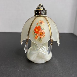 Könnte beinhalten: Eine weiße Glasparfümflasche in Form einer Lampe mit einem Blumendesign auf dem Schirm und kristallartigen Tropfen, die an der Unterseite hängen.