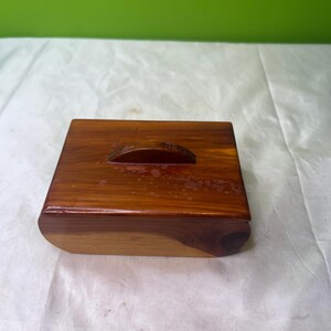 May include: A small, square wooden box with a lid. The box is made of a reddish-brown wood with a curved handle on the lid. The box has rounded edges and a smooth, polished finish.