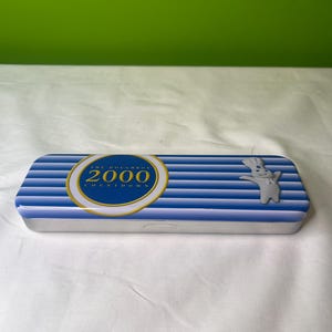Könnte beinhalten: Ein rechteckiges Federmäppchen aus Metall mit blau-weißen Streifen. Das Etui hat ein gold-blaues kreisförmiges Logo mit dem Text "THE DOUGHBOY 2000 COUNTDOWN". Auf der rechten Seite befindet sich eine weiße Doughboy-Figur.