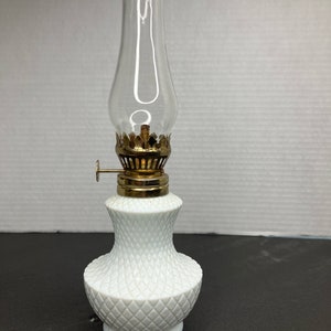 Puede incluir: Una pequeña lámpara de aceite blanca con una chimenea de vidrio transparente. La lámpara tiene una base con diseño de diamantes y una parte superior de metal color latón. La lámpara está diseñada para contener aceite y proporcionar luz. Es un objeto decorativo.